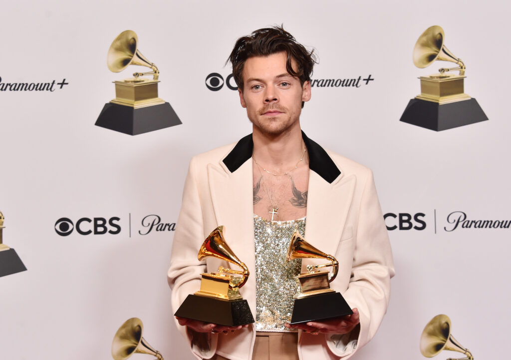 Harry Styles poses in silver top and beige jacket holding two Grammy awards